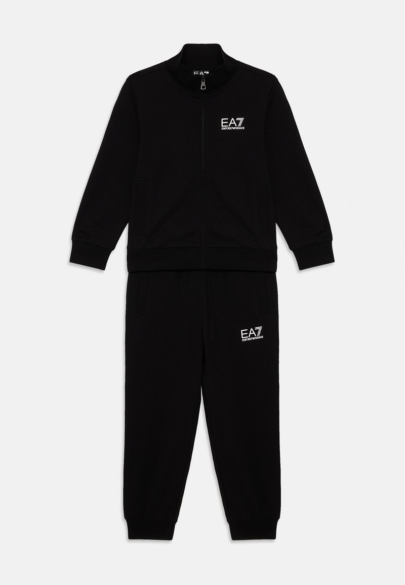Melnā treniņtērps, kas sastāv no ar rāvējslēdzēju aizverama jakas un elastīgas jostas biksēm, abos izstrādājumos ir balts EA7 logo. Mīksta auduma tekstūra.