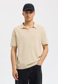Polo shirt beige à manches courtes avec un col V, des poignets et un ourlet côtelés. Le modèle porte une casquette noire et un pantalon sombre, se tenant devant un fond neutre.