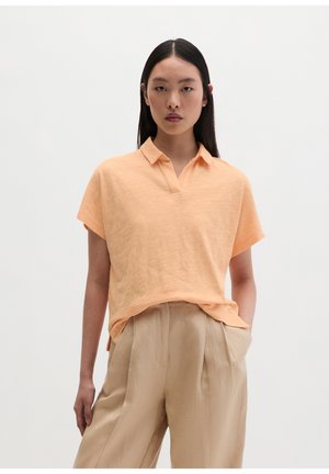 Femme aux longs cheveux foncés portant une chemise pêche à manches courtes avec col et un pantalon beige plissé taille haute, debout avec une main dans la poche.