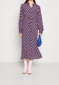 Bedrucktes langärmliges Kleid mit Knopfleiste, tailliertem Gürtel und geometrischem Muster in Rosa, Blau und Lila. Kombiniert mit rosa Stiefeletten.