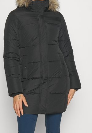 Manteau d’hiver noir matelassé avec fermeture éclair à l’avant et poches pressionnées, capuche bordée de fausse fourrure, porté par une personne avec des tatouages visibles aux mains et un pantalon bleu.