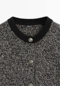 Cardigan tricoté en noir et blanc avec col côtelé noir et trois boutons décoratifs métalliques sur le devant.