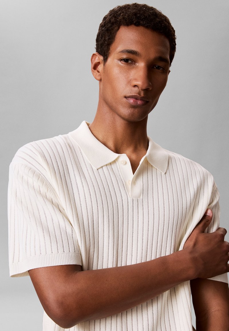 Jeune homme aux cheveux courts et bouclés, portant un polo en tricot blanc à manches courtes, debout, les bras croisés devant un fond gris uni.