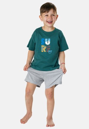 Young boy smiling, wearing a teal t-shirt with colorful "SURF" letters and gray shorts, pulling the shorts slightly outward, barefoot.