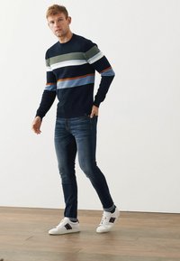 Pulover marin cu dungi verzi, bleo deschis și portocalii. Asortat cu blugi închiși la culoare și adidași albi cu accente colorate. Fundal din podea din lemn.