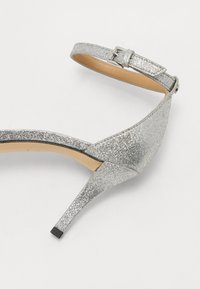 Srebrne štikle s glitterom i tankim remenčićem s kopčom. Imaju elegantan dizajn i špicasti vrh, s crnom blok petom.