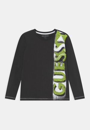 Czarna koszula z długim rękawem, z pionowym, zielonym, popękanym napisem „GUESS” po prawej stronie oraz białym efektem malowania sprayem wokół liter.