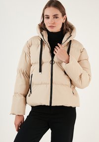 Beige puffer jacket with a hood, featuring a zip front, black accents, and ribbed cuffs. Quilted texture adds volume and warmth.