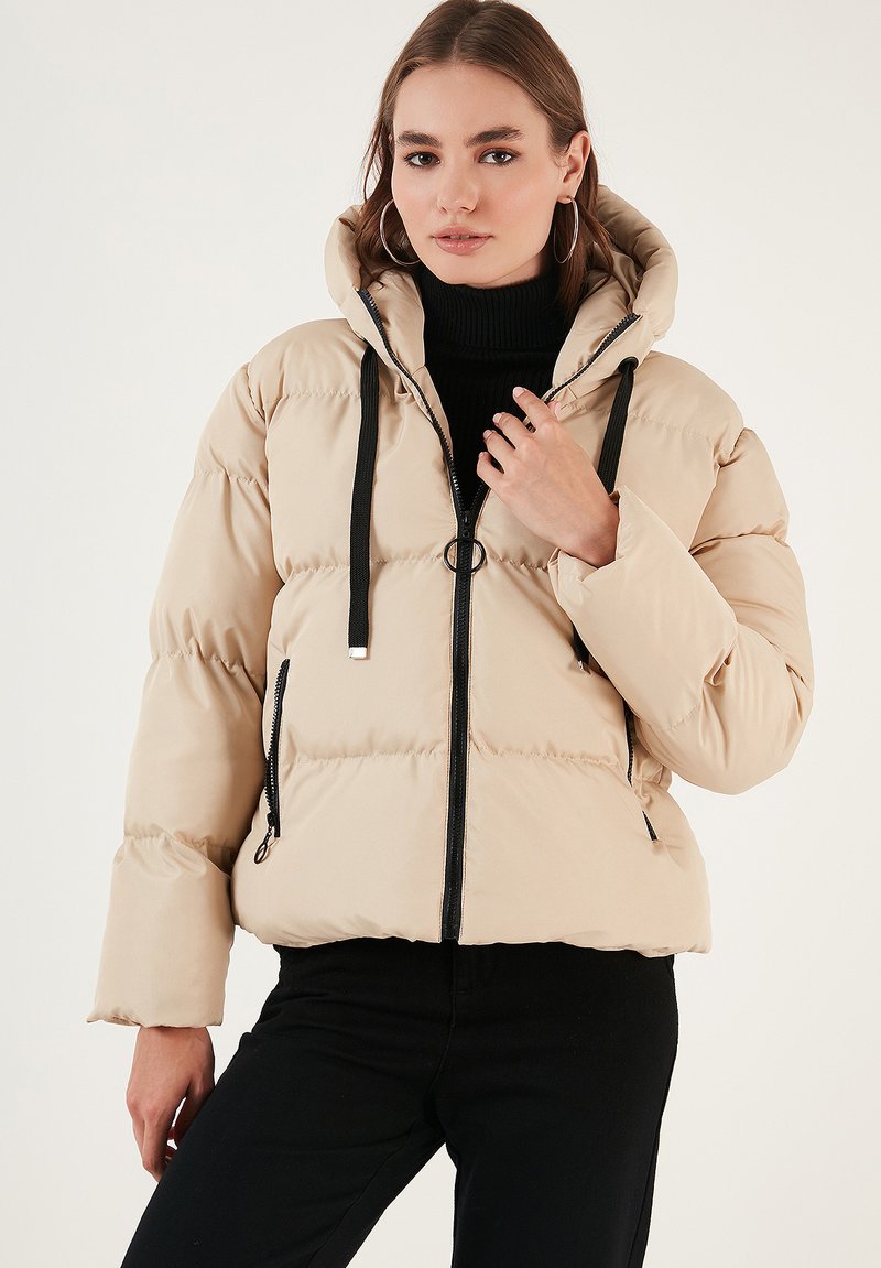 Beige puffer jacket with a hood, featuring a zip front, black accents, and ribbed cuffs. Quilted texture adds volume and warmth.