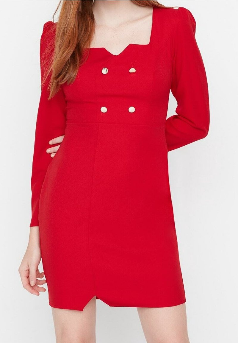 Femme portant une mini-robe rouge à manches longues avec un décolleté carré et quatre boutons dorés sur la poitrine, sur fond blanc.