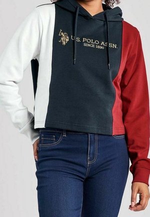 Žena nosí mikinu s barevnými bloky v bílé, námořnické modři a červené barvě s logem „U.S. Polo Assn. Since 1890“, doplněnou tmavě modrými džínami.