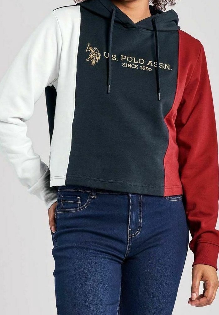 Femme portant un sweat à capuche color-block avec des panneaux blancs, bleu marine et rouges et le logo "U.S. Polo Assn. Since 1890", assorti à un jean bleu foncé.