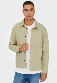 Beige Hemd aus Baumwolle mit Knopfleiste, das zwei Brusttaschen, schwarze Knöpfe und einen klassischen Kragen aufweist, getragen über einem weißen T-Shirt.