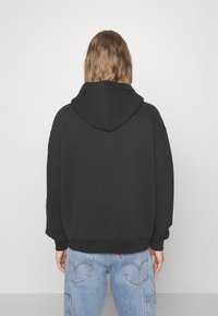 Sudadera negra con una gran capucha, puños y dobladillo acanalados. Corte holgado con una textura suave. Combinada con jeans de mezclilla azul claro.
