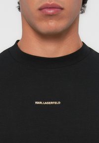 Melnā T-kreklā ar apaļu kakla izgriezumu un mazu zelta logotipu "KARL LAGERFELD" uz krūtīm. Gluda auduma tekstūra, īsas piedurknes.