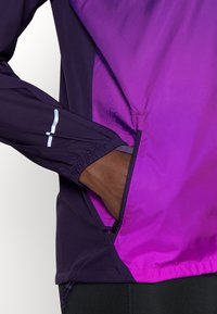 Chaqueta ligera con un degradado que va del púrpura oscuro a un magenta vibrante, con puños elásticos y un bolsillo lateral con cremallera.