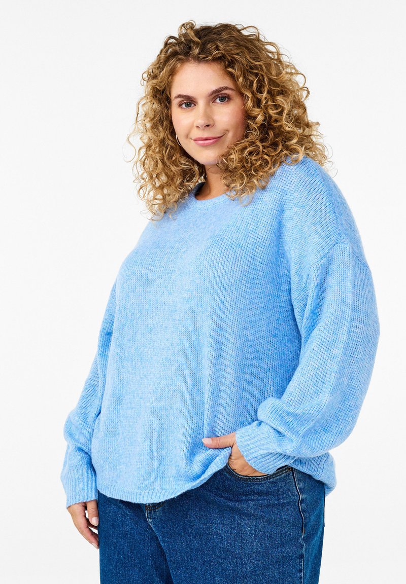 Himmelblauer Strickpullover mit lockerer Passform, überschnittenen Schultern und gerippten Bündchen. Kombiniert mit dunkelblauen Jeans. Strukturiertes Material.