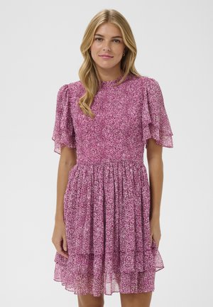 Femme aux longs cheveux blonds portant une robe rose à manches courtes avec un motif et une jupe à volant, debout devant un fond clair uni.