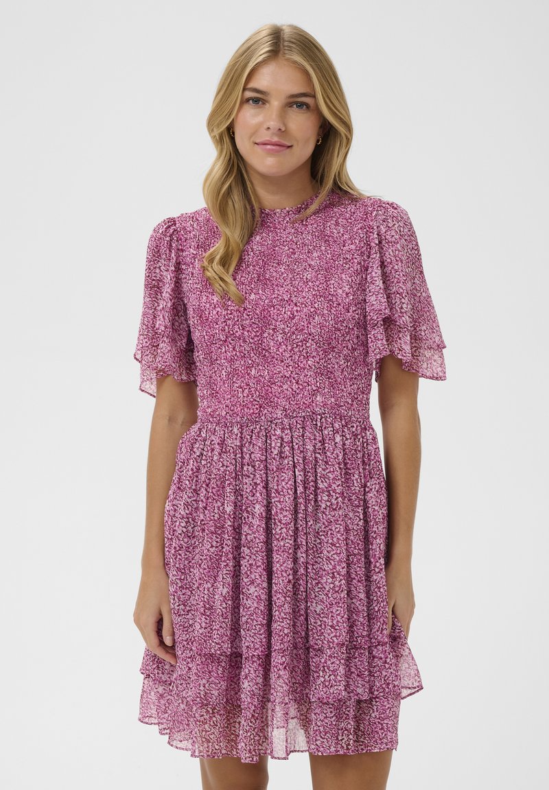 Femme aux longs cheveux blonds portant une robe rose à manches courtes avec un motif et une jupe à volant, debout devant un fond clair uni.