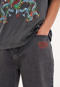 Black graphic t-shirt featuring colorful dragon print; paired with dark denim jeans displaying red embroidered logo on pocket.