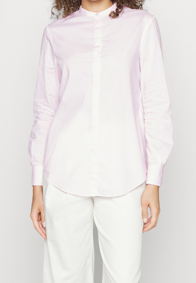 Femme portant une chemise rose pâle à manches longues avec col mandarin et un pantalon blanc, debout devant un fond uni.