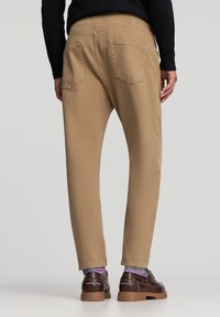 Beżowe spodnie dresowe z elastycznym paskiem w talii, dwoma tylnymi kieszeniami i taperowym krojem. Założone z brązowym obuwiem i wzorzystymi skarpetkami.