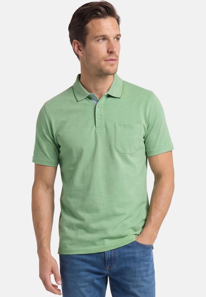 Mann trägt ein hellgrünes, kurzärmeliges Poloshirt mit Brusttasche und blaue Jeans, blickt nach rechts.