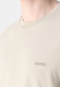 Gros plan d’une personne portant un t-shirt beige à col rond avec le mot « "BOSS" » brodé sur la poitrine.