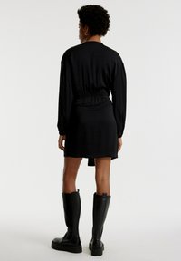 Schwarzes Kleid mit langen Ärmeln und tailliertem Schnitt; strukturierter Stoff; knielang; kombiniert mit knielangen schwarzen Stiefeln.