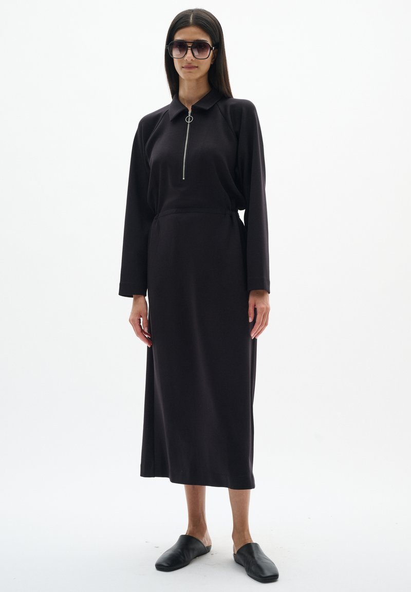 Femme portant une robe midi noire à manches longues avec une fermeture éclair devant, de grandes lunettes de soleil et des chaussures noires sans lacets, debout sur un fond blanc.