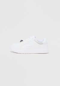 Άσπρο χαμηλό αθλητικό παπούτσι με κορδόνια στο μπροστινό μέρος, ασημί λεπτομέρεια στη φτέρνα και μικρές οπές αερισμού στη πλευρά, πάνω σε απλό φόντο.