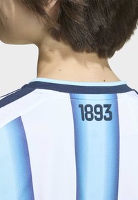 Kratka smešna kosa viđena s leđa, nosi bijelu i plavu prugastu sportsku majicu s natpisom "1893" blizu izreza.