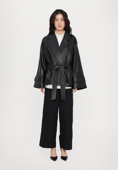 Zwarte leren wikkeljack met wijde mouwen en een geknoopte riem, gedragen over een wit shirt en gecombineerd met zwarte wijde broekspijpen.