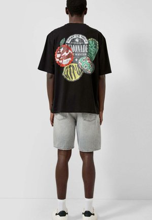 Mann trägt ein schwarzes T-Shirt mit buntem Früchtemotiv und dem Text „Lemonade“ auf dem Rücken, helle Jeansshorts, weiße Socken und Sneakers, steht mit dem Rücken zur Kamera.