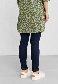 Zelena tunika s uzorkom lišća, uparena s tamnoplavim uskim trapericama i bijelim tenisicama s smeškim naglaskom na leđima.