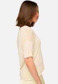 Haut à manches courtes de couleur crème en tissu texturé. Coupe ample avec un ourlet arrondi et des coutures brutes. La vue de côté montre une silhouette décontractée.