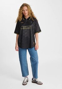 Melnas īsām piedurknēm polokrekls ar apkakli, rakstainu dizainu, dzeltenām detaļām un priekšpuses logotipu. Kombinēts ar zilām taisnu kāju džinsiem.