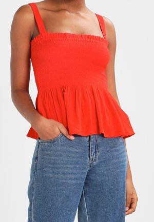 Kvinde iført en rød ærmeløs_top med smock og peplum-ærmekant samt blå højttaljede jeans, den ene hånd i lommen, foran en hvid baggrund.