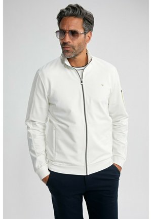 Homme portant une veste blanche zippée, un pantalon foncé et des lunettes de soleil, debout les mains dans les poches devant un fond uni.