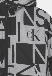 Chaqueta gris y negra con grandes formas geométricas en negrita y el logo "cK" impreso, con cremallera negra y capucha adjunta.