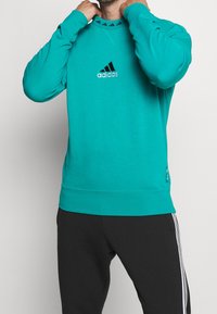 Sweatshirt turquoise avec logo Adidas noir, manches longues, col et poignets côtelés. Présente des détails subtils de design sur le col et l'ourlet.