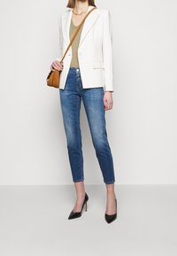 Woman wearing white blazer, beige top, blue cropped jeans, black high heels, and carrying a brown crossbody bag against a plain wall.