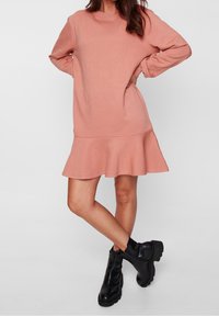 Robe sweat-shirt rose à manches longues, ourlet à volants, texture douce ; associée à des bottines noires à semelle épaisse.