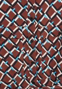 Tissu marron avec un motif géométrique de lignes noires et blanches entrelacées, accentué par de courts coups bleus, présentant des boutons noirs et des poches cousues.