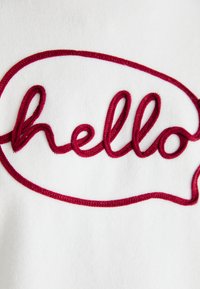 Tissu blanc avec un texte brodé rouge disant "bonjour" à l'intérieur d'une forme de bulle de dialogue. Le texte présente une texture lisse et légèrement en relief.