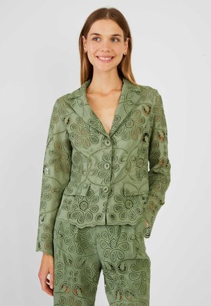 Kobieta ubrana w zielony żakiet z koronką w kwiaty i pasujące spodnie, uśmiechająca się, z jedną ręką w kieszeni, stojąca na jasnym tle.