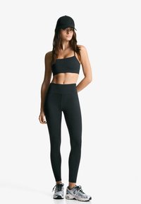 Črn športni crop top in visoko pasne legice iz prožnega materiala, v kombinaciji s srebrnimi teki in črno kapo.