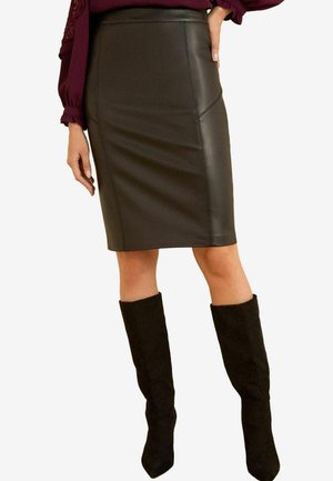 Jupe crayon en cuir noir avec une coupe slim, taille haute et détails de couture visibles. Associée à des bottes noires montantes.