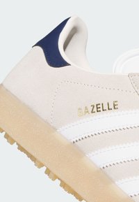 Μπεζ αθλητικό παπούτσι Adidas Gazelle με λευκές ρίγες, χρυσό κείμενο "GAZELLE", σκούρο μπλε ταμπάκι στη φτέρνα και υφή ημιδιάφανης καφέ σόλας.