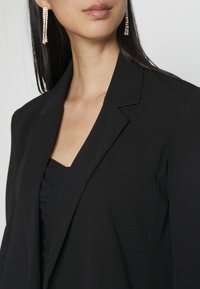 Femme portant un blazer noir et un haut noir avec des boucles d'oreilles pendantes en strass, vue du cou à la poitrine sur un fond uni.